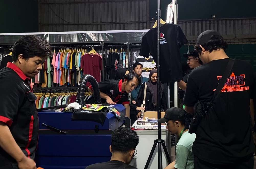 Tim Laysander menunjukkan demo mesin sablon kepada pengunjung dengan berbagai contoh kaos di latar belakang.